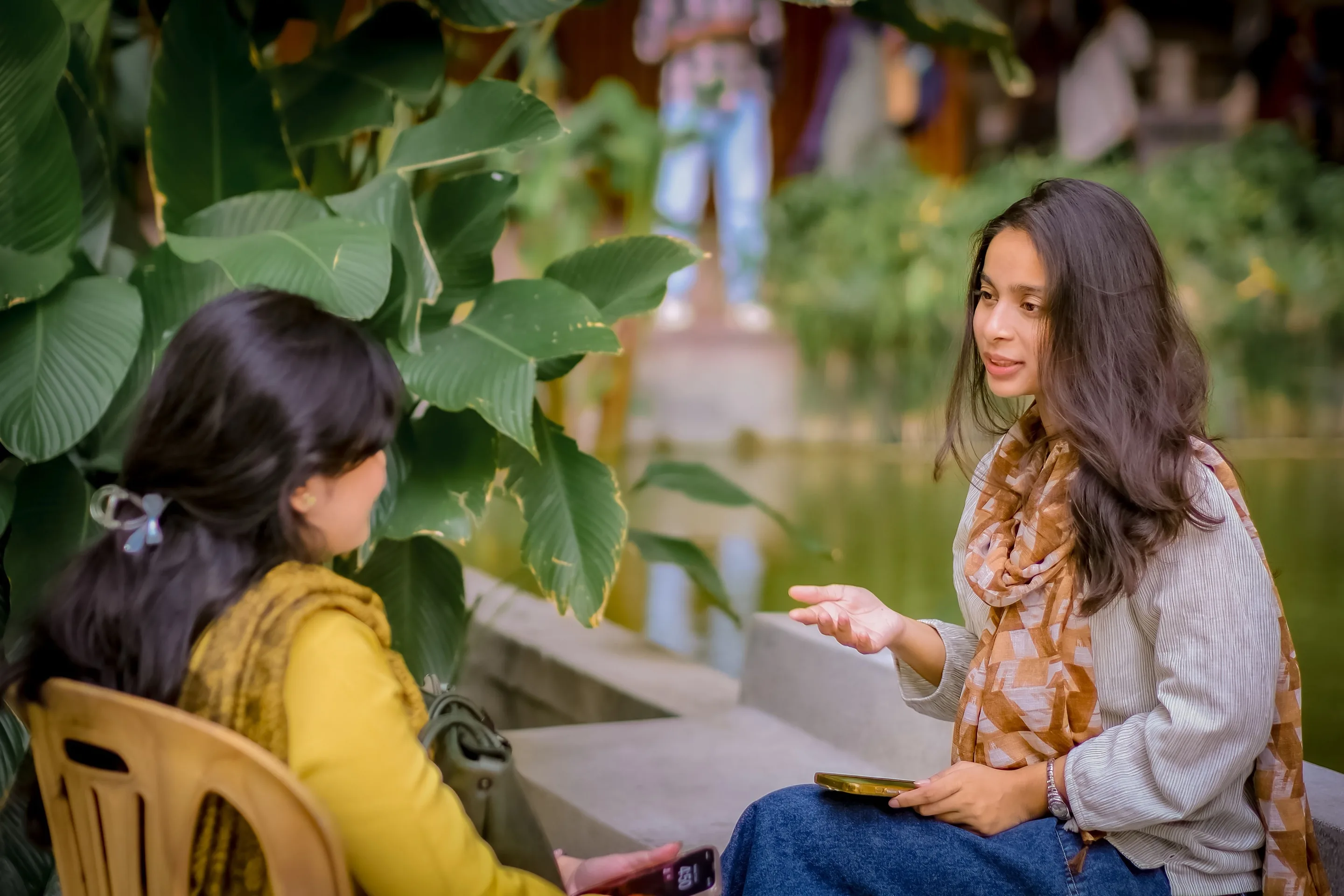 Two women talking outdoors by a pond, one explaining with her hand while the other listens.